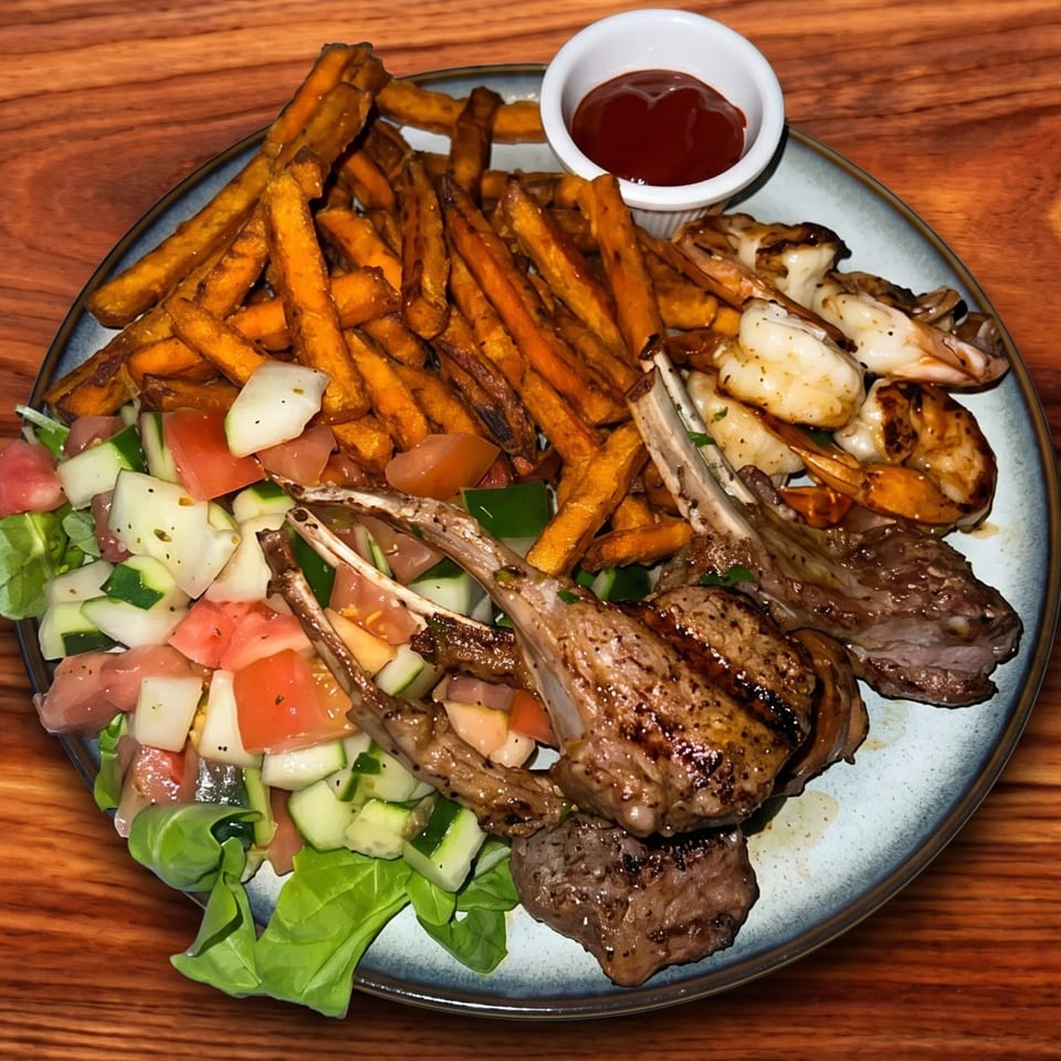 Grilled lamb chops, shrimp, sweet potato fries, and a fresh salad with dressing on a plate.
