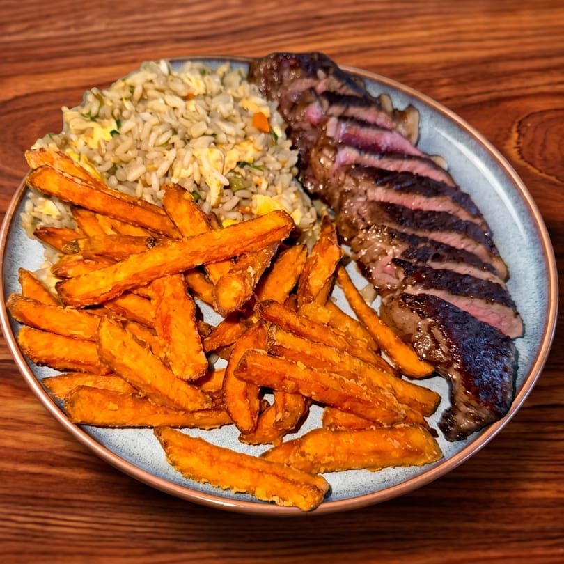 Sliced steak, fried rice, and sweet potato fries on a plate, on a wooden table.