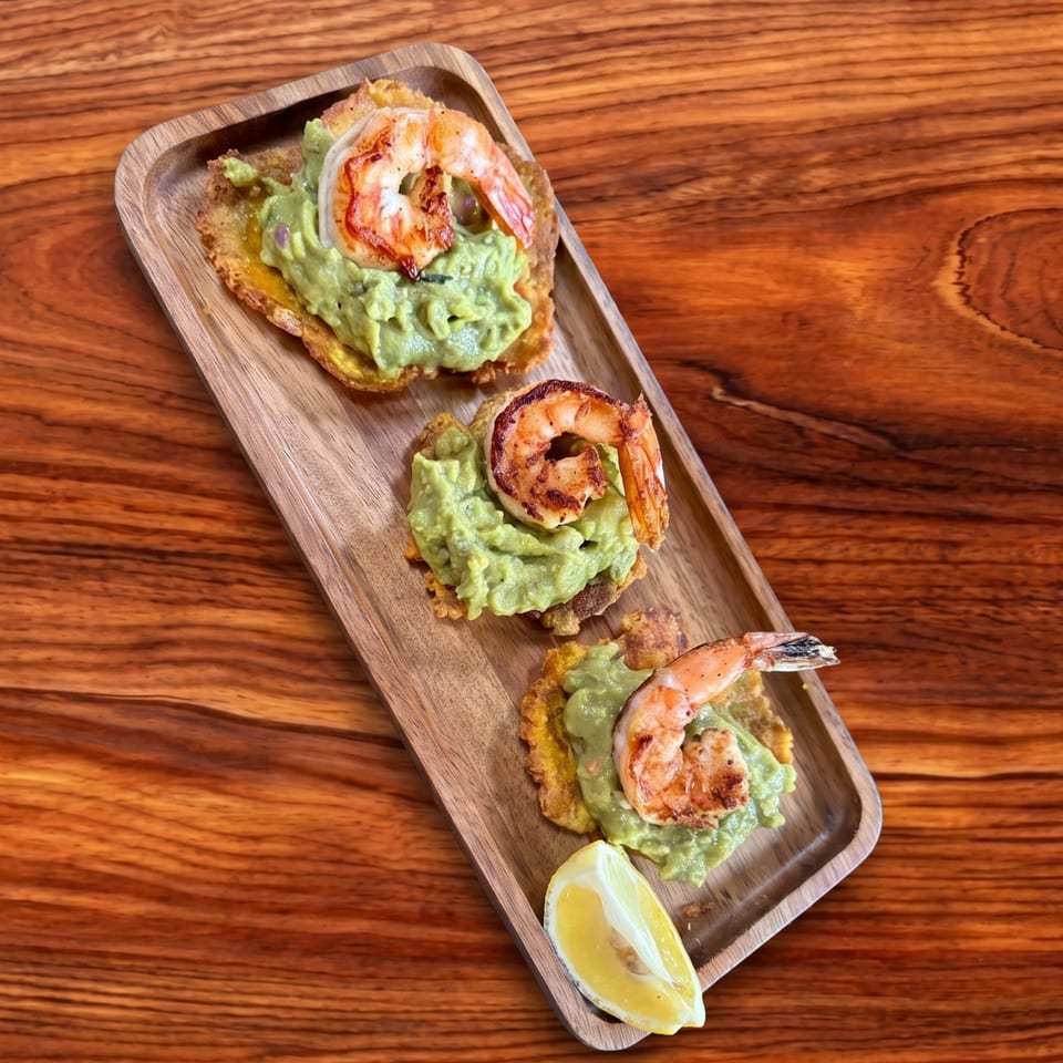 Three shrimp and guacamole tostones with a lemon wedge on a wooden serving tray.