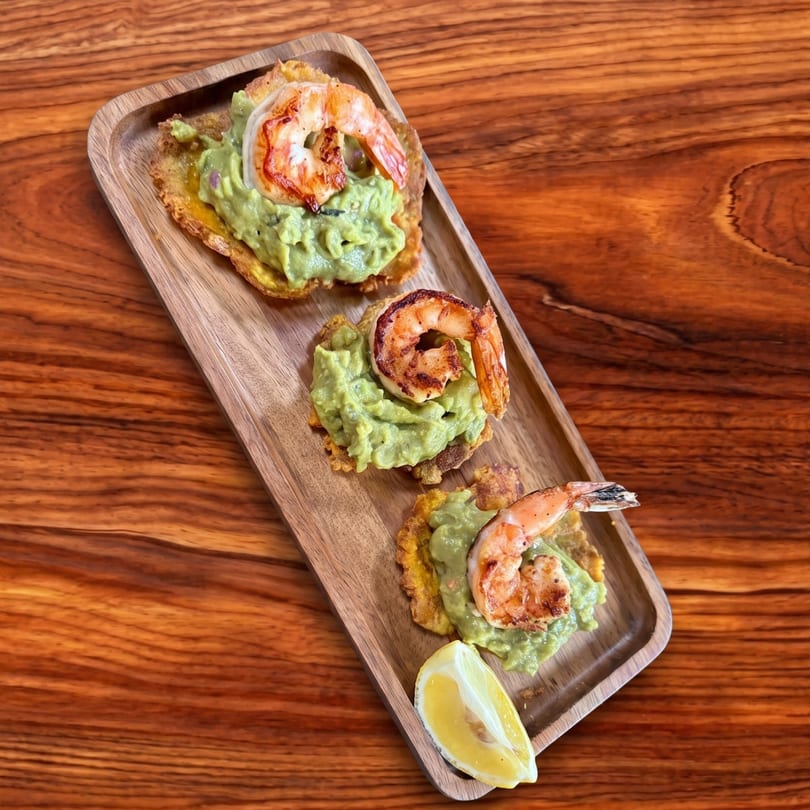 Three shrimp and guacamole tostones with a lemon wedge on a wooden serving tray.